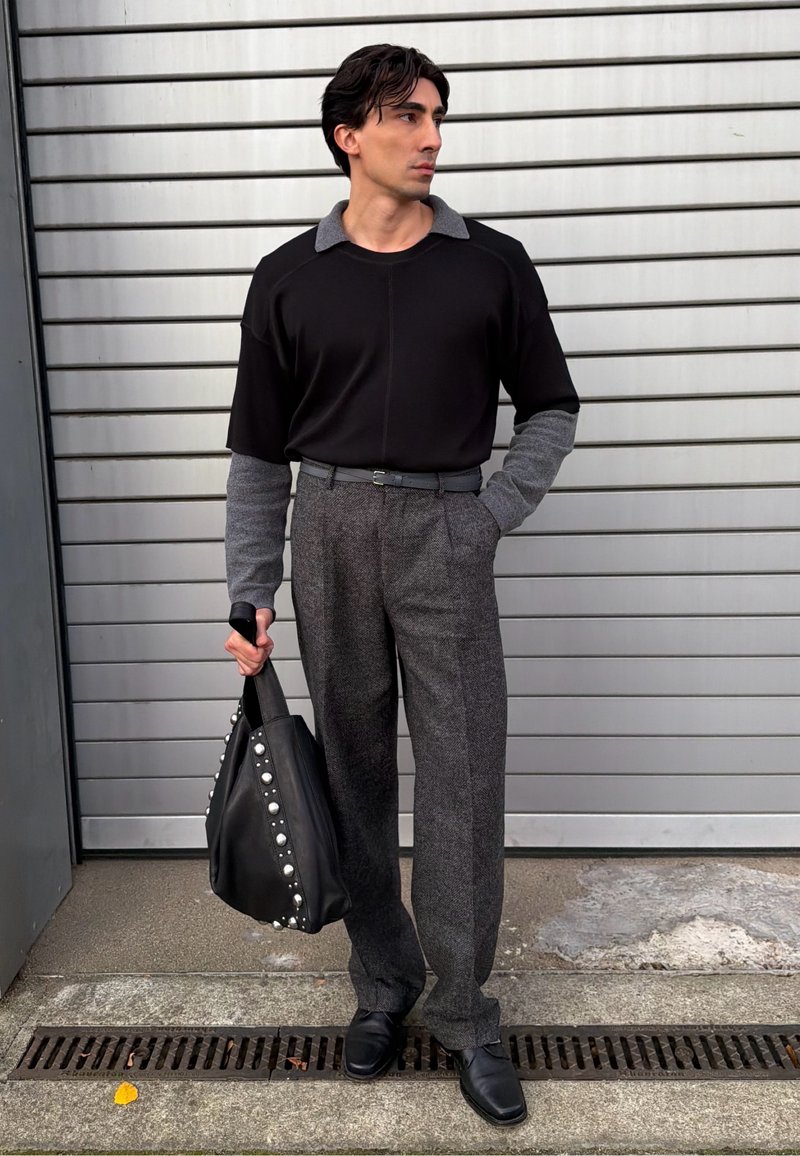 Camicetta nera a maniche corte con accenti grigi, pantaloni grigi su misura, scarpe di pelle nere e una borsa tote nera borchiata. Sfondo texturizzato.