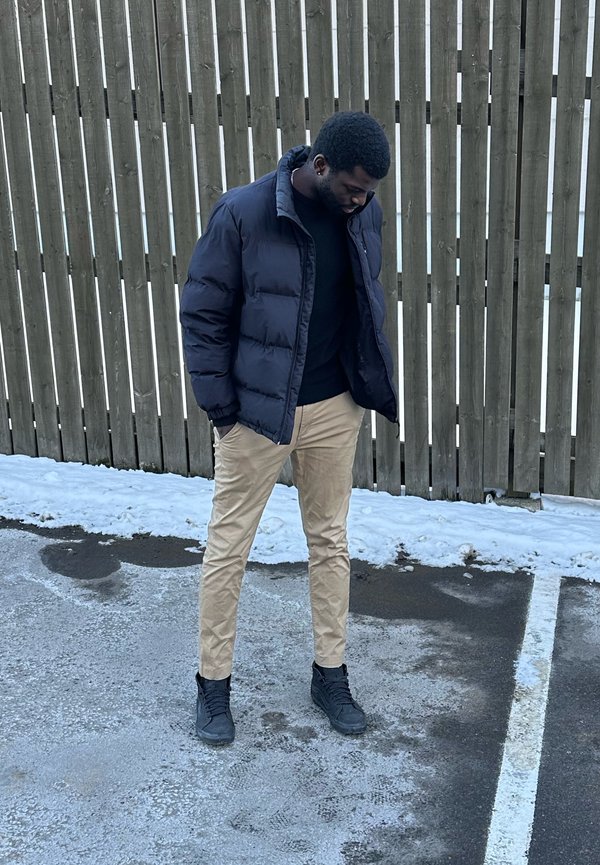 Mężczyzna w czarnej kurtce i beżowych spodniach stoi na zaśnieżonym chodniku przed drewnianym ogrodzeniem, patrząc w dół.