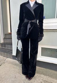 Personne portant un costume en velours noir avec ceinture, chemise blanche, gants noirs, tenant un sac à main noir, debout à l'extérieur près d'un escalier carrelé et d'une fenêtre.