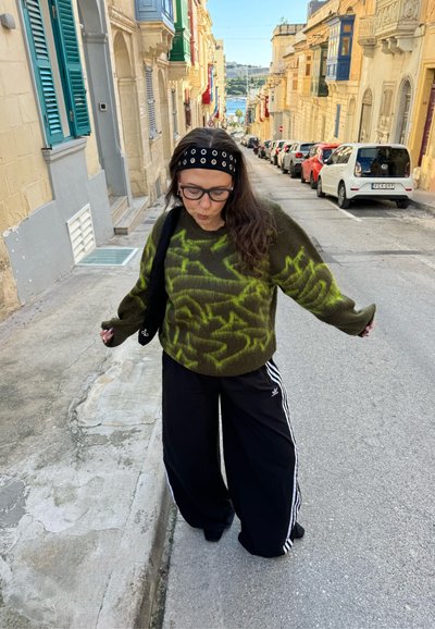 Mujer joven con gafas, diadema y suéter verde con estampado, de pie en una calle estrecha bordeada de coches aparcados y balcones coloridos.