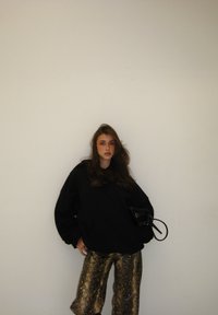 Sweat à capuche noir surdimensionné à manches longues, associé à un pantalon large imprimé serpent brillant en or et marron. Tenant un sac à main noir.