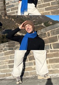 Črna majica z dolgimi rokavi, modri šal z resicami, rjava pletena kapa; svetlo bež široke hlače, sive superge. V ozadju Veliki zid.