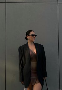 Blazer oversize a righe nere indossato sopra un vestito marrone con volant. Gli accessori includono occhiali da sole piccoli e orecchini grandi. Sfondo grigio.