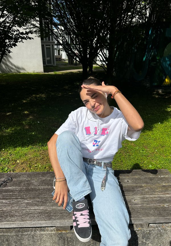 Witte T-shirt met roze en blauwe grafische print, lichtblauwe jeans, zwarte sneakers met roze accenten, zittend op een houten bank buiten.