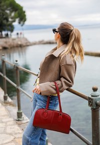 Sac à main en cuir rouge avec une forme structurée et deux poignées supérieures, associé à une veste en laine beige et un jean bleu, devant un décor aquatique.