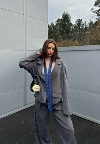 Jeune femme portant un costume gris surdimensionné et une cravate bleue à motifs, posant en extérieur devant un mur gris avec des arbres en arrière-plan.