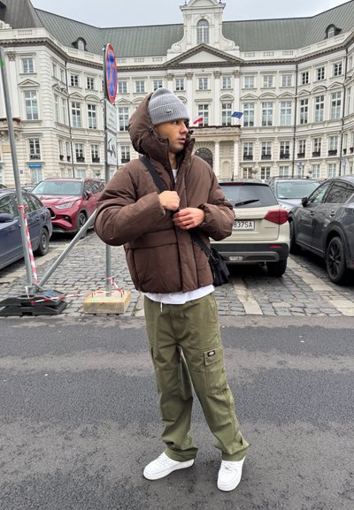Chaqueta de plumas marrón con capucha, camiseta blanca, pantalones cargo verdes oliva y zapatillas blancas. El fondo presenta un entorno urbano con vehículos.