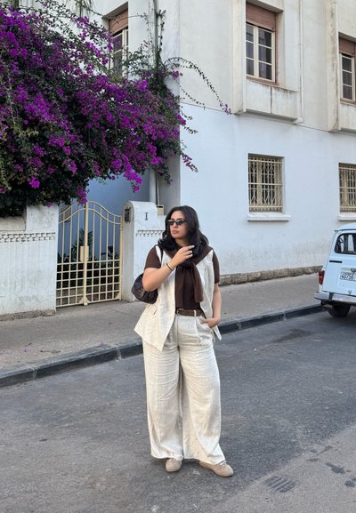 Mujer con conjunto de lino beige y gafas de sol, de pie en la calle cerca de un edificio blanco con una enredadera de flores moradas y un coche antiguo estacionado.