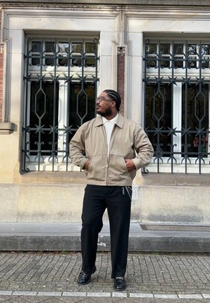 Chaqueta bomber beige, camisa blanca, pantalones negros y zapatos negros brillantes. Cabello a rayas, de pie frente a ventanas ornamentadas con rejas de metal.