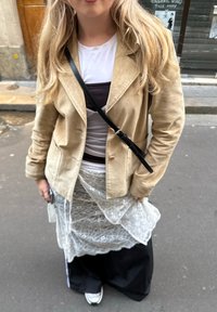 Béžová semišová bunda přes vrstvený outfit se bílým topem, hnědou spodní košilí, průsvitnou krajkovou sukní a černými širokými kalhotami. Černá kabelka přes rameno.