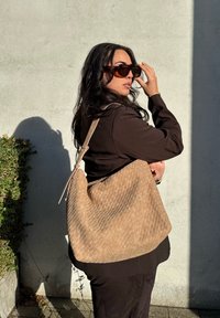 Vævet tan skuldertaske med en buet form, der har en glat læderrem og en struktureret overflade. Modellen bærer mørkt tøj og solbriller.