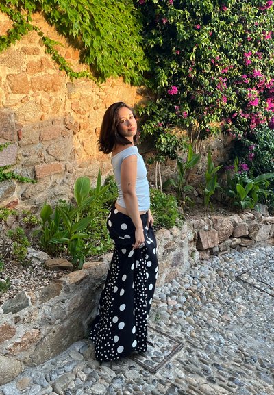 Top azul claro sin mangas, pantalones anchos negros con grandes lunares blancos, de pie en un camino de adoquines cerca de plantas y flores verdes.