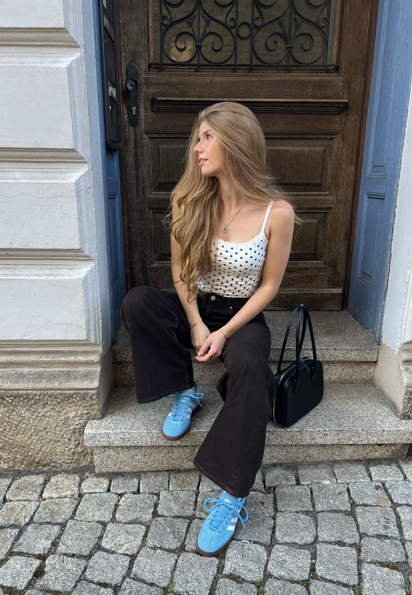 Młoda kobieta z długimi blond włosami siedzi na kamiennych schodach w niebieskich trampkach, czarnych spodniach i białym topie w groszki, obok czarnej torebki przy drewnianych drzwiach.