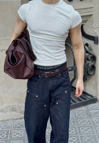 Hvid tætsiddende t-shirt, mørke denimjeans med nitte detaljer, brunt bælte og en bordeaux lædertaske med blød tekstur, båret under armen.