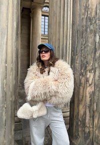 Femme portant un manteau beige duveteux, un pantalon de survêtement gris, des lunettes de soleil et une casquette bleue, debout les bras croisés entre de grandes colonnes en pierre.