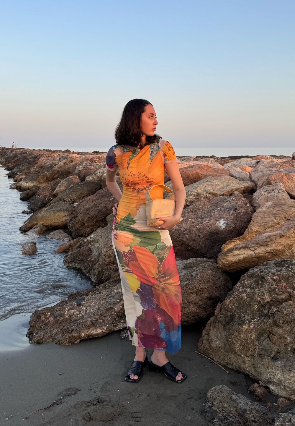 Abito con fantasia floreale caratterizzato da colori vivaci (arancione, rosso, verde) e un busto aderente, che scende fino alle caviglie. La modella tiene in mano una piccola borsa testurizzata.