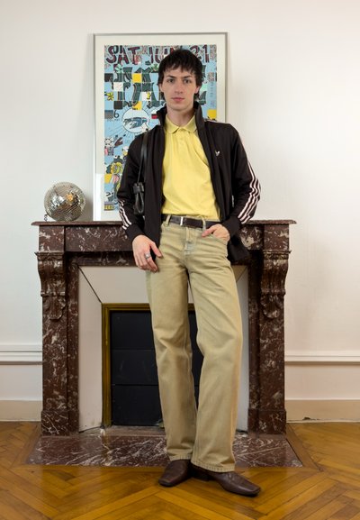 Joven con polo amarillo y pantalones beige se apoya en una chimenea de mármol marrón, con una bola de discoteca y obras de arte coloridas enmarcadas detrás.
