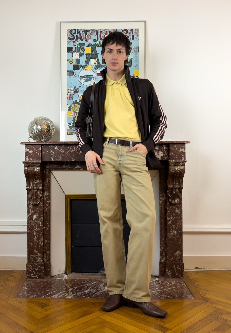 Jeune personne en polo jaune et pantalon beige appuyée sur une cheminée en marbre marron, avec une boule disco et des œuvres d'art encadrées colorées en arrière-plan.