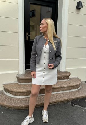 Chaqueta gris corta sobre un vestido blanco con detalles de botones, combinada con zapatillas de color claro. Los escalones y una puerta oscura ofrecen contexto.