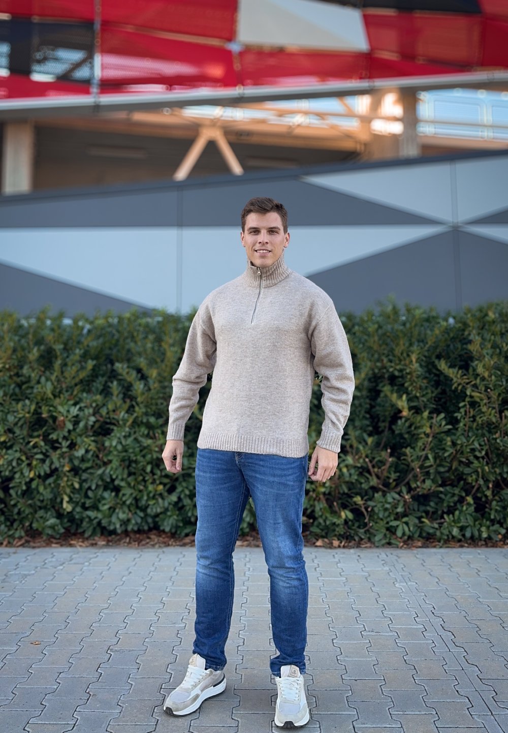 Μπεζ πλεκτός πούλος με ψηλό γιακά και φερμουάρ, εφαρμοστές μανσέτες, συνδυασμένος με μπλε τζιν και ανοιχτό γκρι αθλητικά παπούτσια. Πράσινα φυλλώματα στο φόντο.