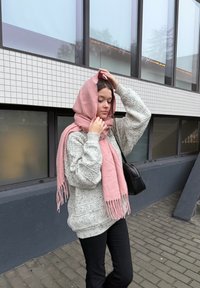 Világos rózsaszín sál a fején, texturált szürke pulóver, fekete nadrág, épület nagy ablakai és csempézett fala mellett állva a szabadban.