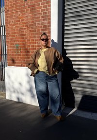 Brun bomberjakke over en gulstripet skjorte, kombinert med løse blå jeans og brune sko. Tilbehøret inkluderer sorte solbriller og en teksturert svart veske.