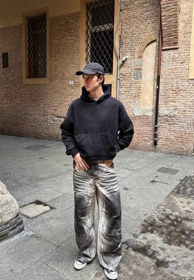 Sudadera con capucha negra, pantalones grises anchos y desgastados con manchas negras, gorra negra y zapatillas blancas, de pie en una calle de piedra.