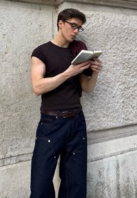 Czarny dopasowany t-shirt, ciemnoniebieskie szerokie spodnie dżinsowe z metalowymi akcentami, brązowy pasek, czytający książkę, na tle teksturowanej kamiennej ściany.
