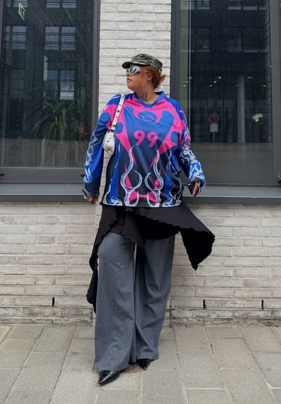 Persona que lleva una camisa de manga larga colorida con el número 99, pantalones anchos grises, zapatos negros puntiagudos, gafas de sol y una gorra militar, de pie junto a una ventana.