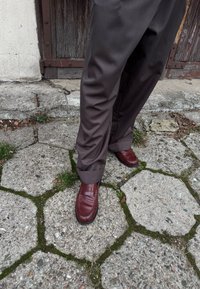 Brūnas bikses ar gludu tekstūru, kopā ar bordo ādas loafers kurpēm ar šūtu akcentu. Apkārtne ietver nevienmērīgu akmens bruģi.