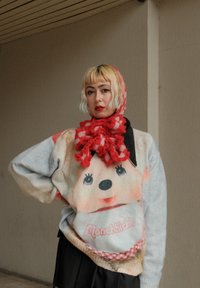 Maglione lavorato a maglia con un design della faccia di Monchhichi in morbidi colori pastello, con un colletto nero e una sciarpa a quadri rossi, creando un look giocoso.