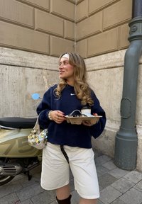 Felpa oversize blu navy con logo Nike bianco, shorts bianchi, una cintura nera a fantasia e una borsa tempestata di paillettes. Tiene una bevanda in un vassoio di cartone.