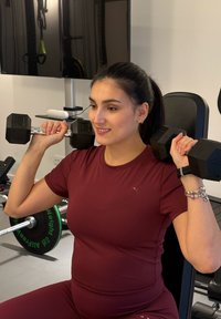 Femme en tenue de sport bordeaux soulevant des haltères de 5 kg assise dans une salle de sport à domicile avec des poids et une télévision en arrière-plan.