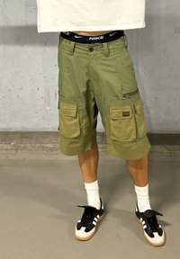 Olijfgroene cargoshorts met meerdere zakken, beige accenten en een Nike tailleband. Gecombineerd met zwart-witte sneakers en witte sokken.