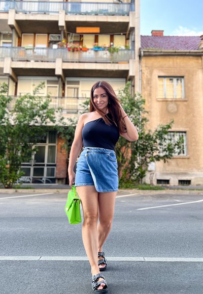 Top negro de un solo hombro, falda envolvente de denim, bolso verde y sandalias negras con tachuelas. Fondo urbano con balcones y vegetación.