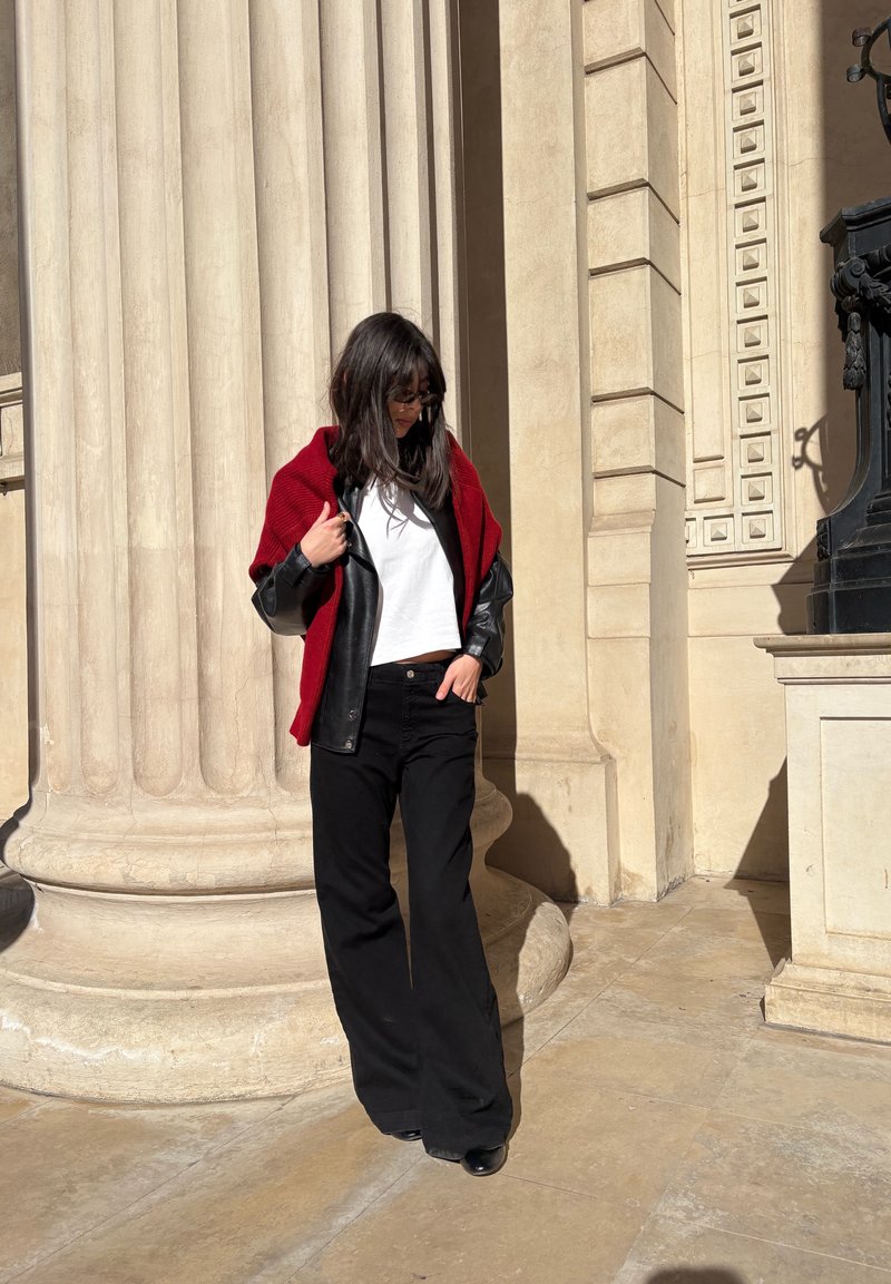 Femme portant une veste en cuir noire, un haut blanc, un châle rouge et un pantalon large noir, debout près de hautes colonnes en pierre, sous le soleil.