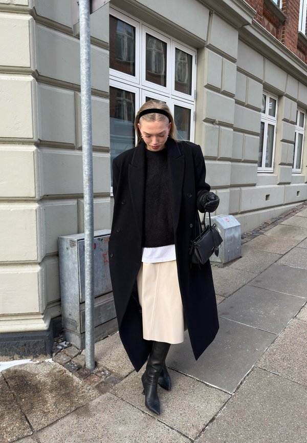 Kobieta ubrana w czarny płaszcz, czarny sweter, beżową spódnicę, czarne kozaki i rękawiczki stoi na chodniku obok budynku z dużymi oknami.