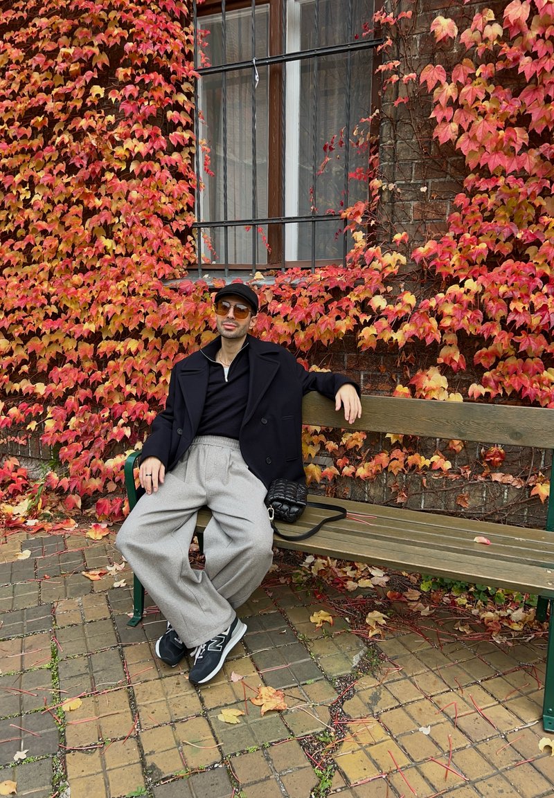 Blazer preto com cauda sobre uma camisa preta, calças largas cinzas, sapatos pretos e um boné preto, sentado num banco de madeira entre folhagem vermelha.