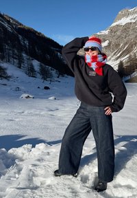 Personne portant un chapeau rouge et bleu et une écharpe, un sweat-shirt noir et un jean, debout sur un sol enneigé avec des montagnes et un ciel dégagé en arrière-plan.