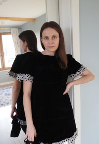 Robe en velours noir avec manches courtes bouffantes, ornée d'un volant à pois. Texture lisse, longueur au genou, et coupe évasée.