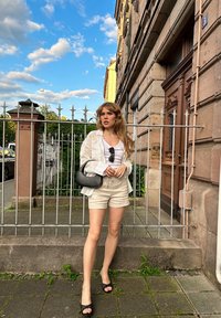 Camicia bianca leggera con bottoni sopra un top bianco, pantaloni corti beige, borsa nera a tracolla, sandali neri con lacci, in piedi accanto a una recinzione metallica.