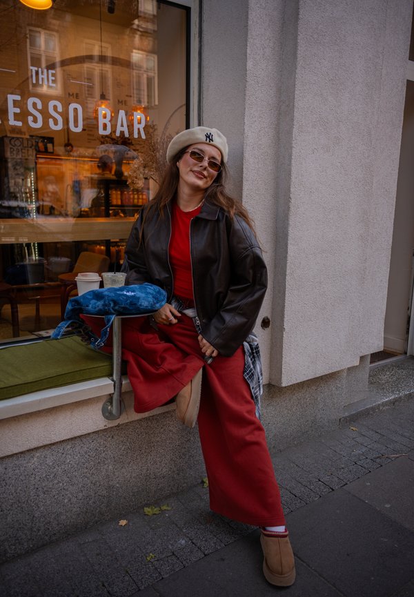 Una mujer con boina, gafas de sol, chaqueta de cuero y ropa roja posa junto al escaparate de una cafetería; en el alféizar hay un bolso azul.