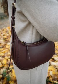 Naine kannab beeži veste ja hoiab pruunist nahast õlakotti, millel on lukku, seistes sügislehtede peal õues.