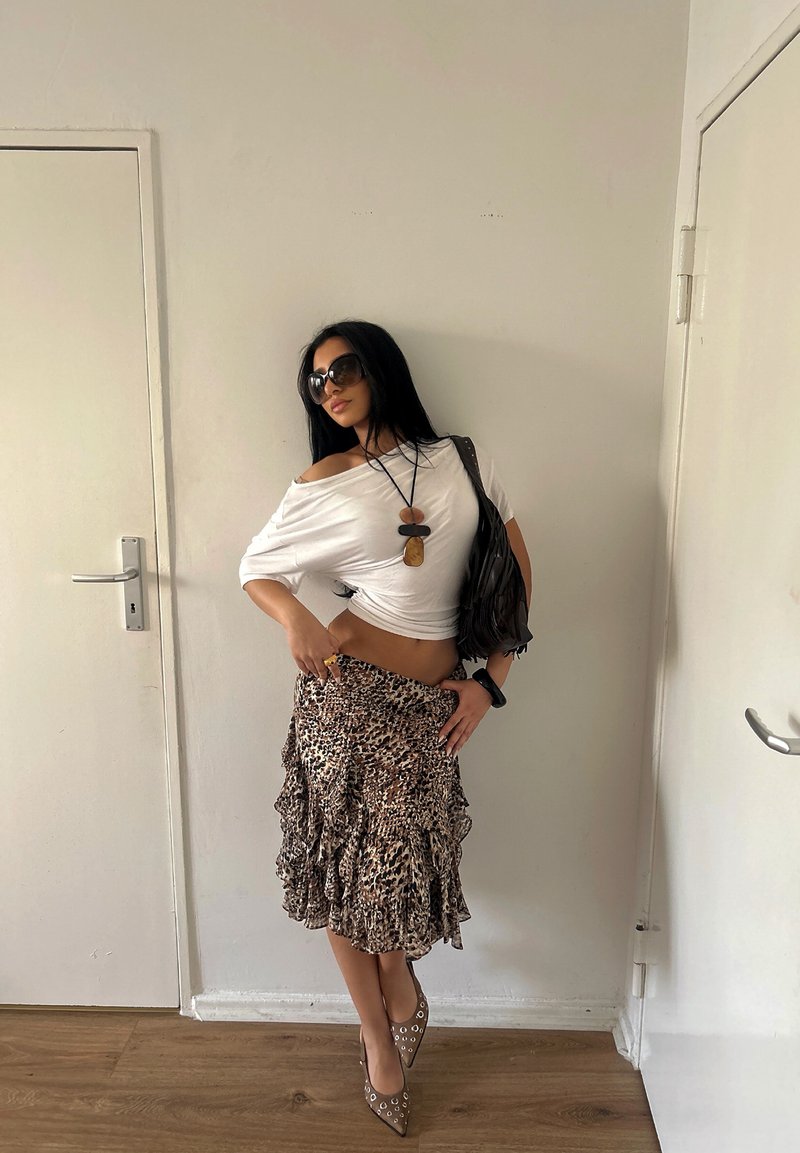 Femme portant un haut blanc à épaules dénudées, une jupe imprimé léopard, des lunettes de soleil et des chaussures pointues, debout à l'intérieur entre deux portes blanches fermées.