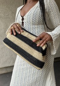 Πλεκτή τσάντα clutch με εναλλασσόμενες μαύρες και μπεζ ρίγες, σε ορθογώνιο σχήμα και υφή, κρατημένη στα χέρια σε φωτεινό φόντο.