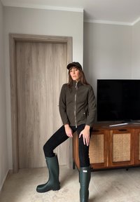 Γυναίκα με σκούρο πράσινο σακάκι, μαύρο παντελόνι, πράσινες μπότες μέχρι το γόνατο και καπέλο, ακουμπά σε ξύλινη ντουλάπα με τηλεόραση σε δωμάτιο με ουδέτερους τόνους.