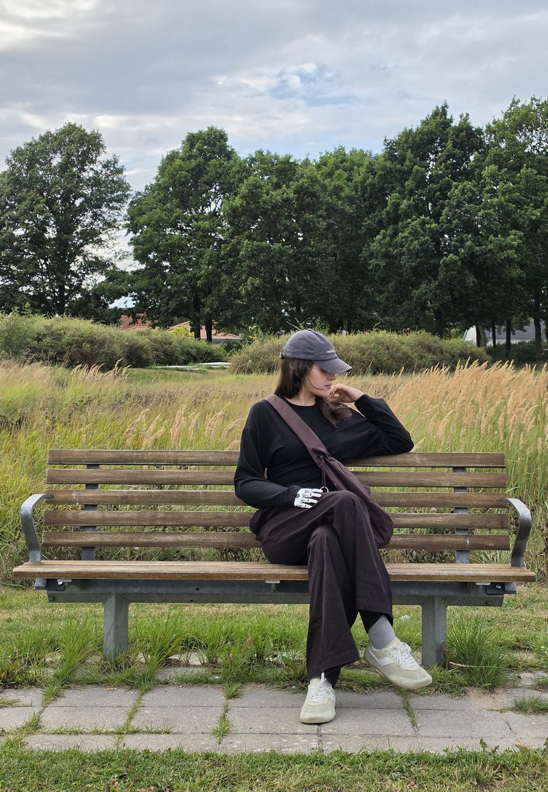 Personne portant des vêtements foncés et une casquette assise sur un banc en bois dans un parc avec de hautes herbes et des arbres en arrière-plan.