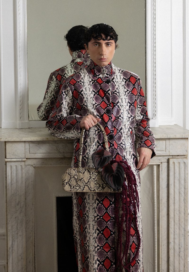 Personne portant un manteau à motif serpent rouge, noir et blanc se tient devant une cheminée en marbre, tenant un sac à main assorti au motif serpent avec des fleurs sombres.