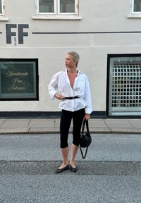 Λευκό υπερμεγέθες πουκάμισο με κουμπιά, μαύρη ζώνη, μαύρες κοντές κολάν, μαύρη τσάντα και μαύρα παπούτσια με μαύρες πουά; ομαλή, χαλαρή εμφάνιση.
