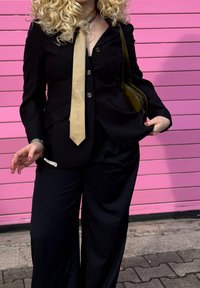 Juodas pritaikytas švarkas su auksiniais mygtukais, smėlio spalvos kaklaraištis, laisvai krentančios juodos kelnės ir alyvuogių žalios spalvos krepšys. Fone matyti rožinės horizontalios linijos.